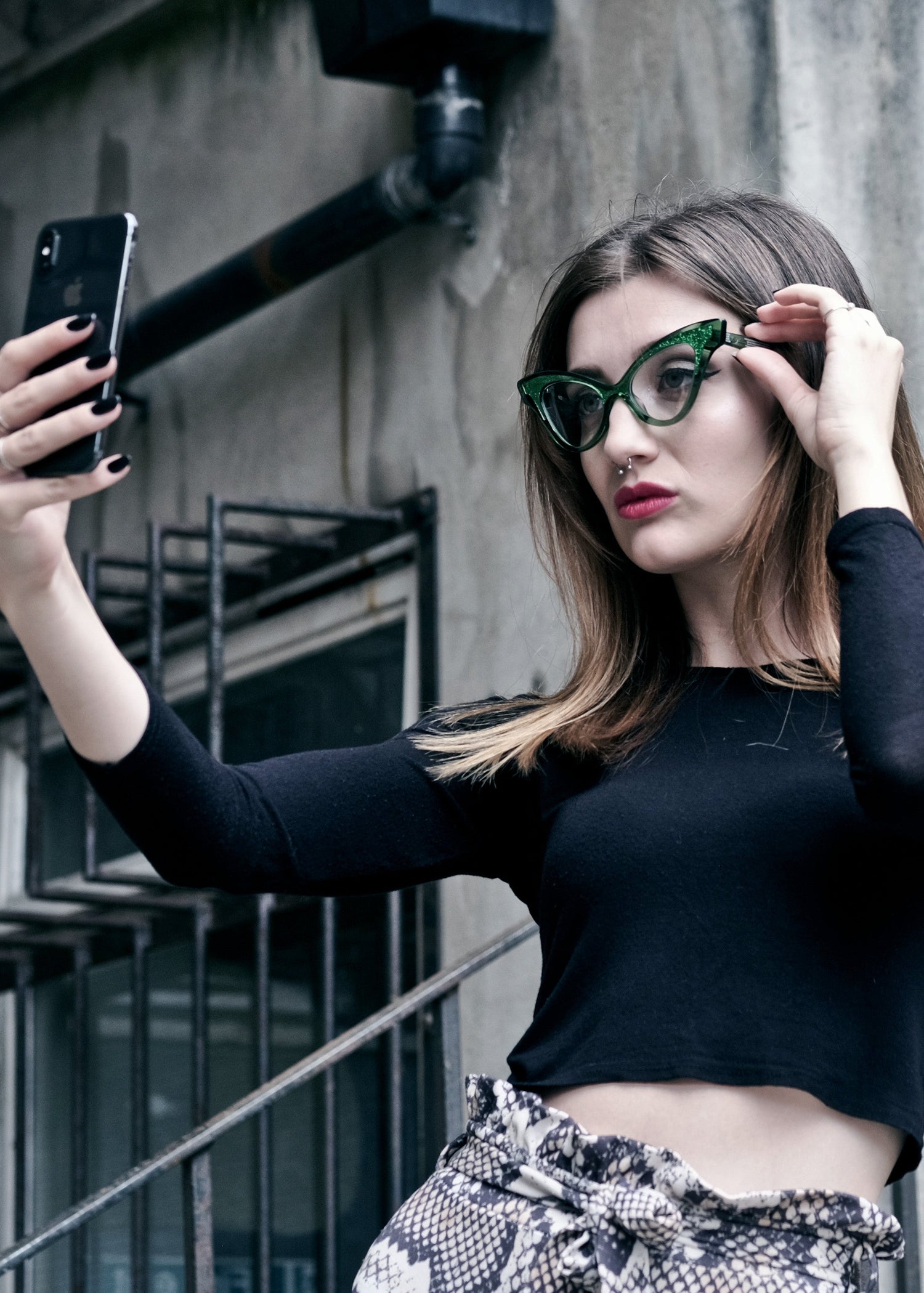 Woman taking a selfie outdoors against a concrete wall.  Wearing green cat eye glasses.